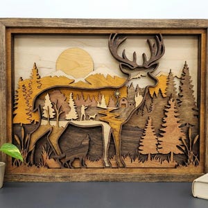 Op de afbeelding: Houten gelaagd kunstwerk dat een hert in een bosscene afbeeldt. Het kunstwerk toont een silhouet van een hert, bomen, bergen en een zon, allemaal gemaakt van verschillende houttinten. Het is ingelijst in een donkerbruine houten lijst.