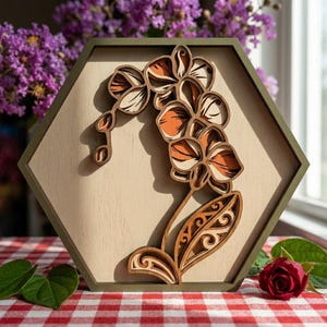 Może przedstawiać: Drewniane, warstwowe dzieło sztuki przedstawiające kwiat orchidei. Kwiat jest pomarańczowy, kremowy i brązowy, na jasnobeżowym tle w zielonej, sześciokątnej ramie. Grafika jest prezentowana na czerwono-białej kraciastej serwetce.