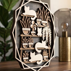 May include: Wooden wall art featuring a layered design of cats and potted plants. The artwork has a dark gray background with light brown shelves and cat silhouettes. The outer frame is a light brown geometric shape.