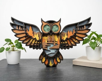 May include: A multi-layered wooden owl wall art piece featuring outstretched wings. The design incorporates a forest landscape with orange and yellow trees, a blue river, and dark brown outlines. The owl has green eyes with a hexagonal pattern.