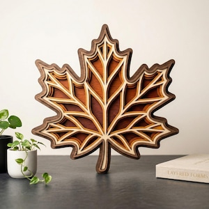 May include: A layered wooden maple leaf sculpture in shades of brown and tan. The leaf is intricately cut with multiple layers, creating a three-dimensional effect. The sculpture is displayed on a dark surface, with small potted plants and a book nearby.