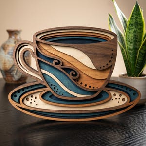 Könnte beinhalten: Eine mehrschichtige Holztasse und Untertasse. Die Tasse hat einen dunkelbraunen Rand mit Schichten aus Blau, Beige und hellem Holz. Die Untertasse hat ein passendes Design mit einem dunkelblauen Rand und kleinen ausgeschnittenen Kreisen. Ein dekoratives Stück für das Zuhause.