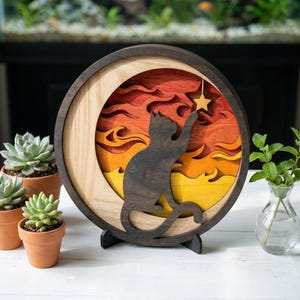 Könnte beinhalten: Runde Wandkunst aus Holz mit einer Katze, die nach einem Stern greift. Das Kunstwerk hat ein mehrschichtiges Design mit warmen Farben wie Orange, Gelb und Braun. Die Katzen-Silhouette ist dunkelbraun, und das Stück wird von einem schwarzen Ständer getragen.