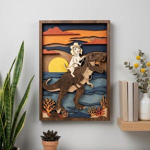 Op de afbeelding: Gelaagd houten kunstwerk met een hond met cowboyhoed die een Tyrannosaurus Rex berijdt. De scène toont een zonsondergang met een gele zon, blauw water en een oranje en blauwe lucht. De lijst is donkerbruin.