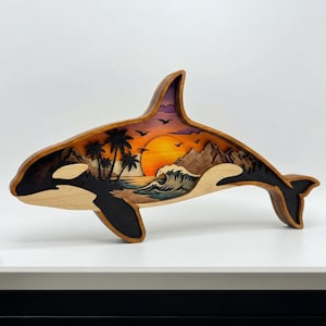 May include: Wooden orca sculpture with a layered design featuring a sunset over a beach scene. The orca is primarily light wood with black accents, and the interior scene includes palm trees, mountains, and a vibrant orange sun.
