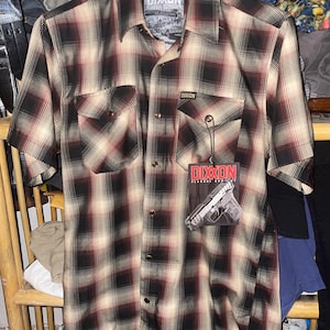 May include: A short-sleeved plaid shirt in black, cream, and burgundy. The shirt has two chest pockets and a button-down front. A tag with the brand name "DIXXON" and a graphic of a handgun is attached.