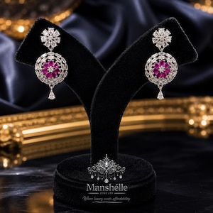 Op de afbeelding: Elegante zilveren oorbellen met een bloemmotief. Elke oorbel heeft een cluster van roze edelstenen in het midden, omringd door heldere stenen. Een druppelvormige edelsteen hangt eronder. De oorbellen worden op een zwarte standaard gepresenteerd.
