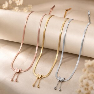 Può includere: Tre collane a catena piatta a serpente nei toni dell'oro rosa, oro giallo e argento. Ogni collana presenta un ciondolo a farfalla centrale con due delicate catenelle pendenti che terminano in piccole sfere e una catena regolabile.