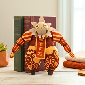 May include: A plush doll with a sun-like head and a patterned outfit in shades of red, yellow, and brown. The doll has a white flower-shaped crown and a banner with text. The doll is standing in front of books and a succulent plant.