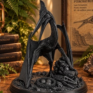 Op de afbeelding: Een zwart Pterosaurus-beeldje op een ronde voet met een nest met drie eieren en een skeletrest. Het wezen heeft een lange snavel, een puntige kam en uitgestrekte vleugels, uitgevoerd in een effen matzwarte afwerking.
