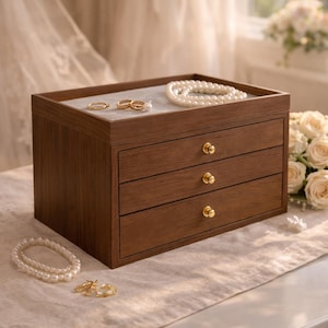 Puede incluir: Una caja de joyería de madera con tres cajones y un compartimento superior. La caja es de color marrón cálido con pomos dorados. El compartimento superior contiene anillos y un collar de perlas. Un collar de perlas y anillos están sobre la mesa.