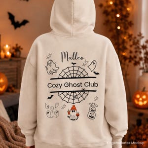 Könnte beinhalten: Ein beigefarbener Kapuzenpullover mit einer schwarzen Grafik auf der Rückseite, die den Text "Matteo" und "Cozy Ghost Club" zeigt. Das Design enthält Illustrationen von Geistern, Spinnennetzen, Fledermäusen, Sternen und einem Geist mit orangefarbener Mütze.