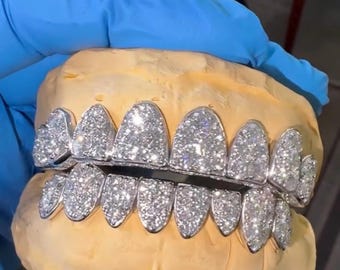 May include: A set of custom-made, silver-colored grillz encrusted with numerous sparkling, clear gemstones. The grillz are displayed on a light-yellow dental mold, showcasing the intricate design and craftsmanship. The overall aesthetic is luxurious and eye-catching.