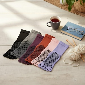 Puede incluir: Una variedad de calcetines de dedos en varios colores, incluyendo negro, gris, morado y rosa, dispuestos sobre una superficie de madera clara. Cada calcetín presenta un patrón antideslizante en la suela. También hay una taza de té, un libro y utensilios de tejido.