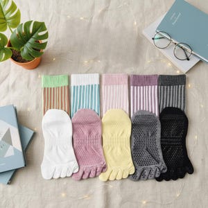 Op de afbeelding: Een verzameling teensokken in diverse kleuren, waaronder wit, roze, geel, grijs en zwart. Elke paar heeft een gestreept patroon op het bovenste gedeelte. De sokken zijn ontworpen met individuele teengleuven.