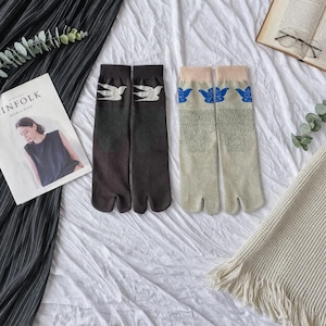 Puede incluir: Dos pares de calcetines con puntera dividida. Un par es marrón oscuro con diseños de pájaros blancos, el otro es beige claro con diseños de pájaros azules. Los calcetines se muestran sobre una superficie blanca con una revista y un libro en el fondo.