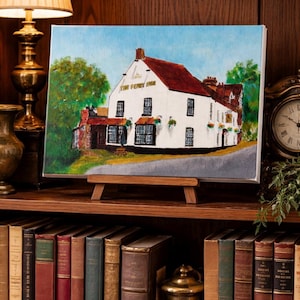 May include: An oil painting of a white building with a red roof, titled "The Ferry Inn", displayed on a wooden easel. The painting is set against a backdrop of a bookshelf, a lamp, and a clock.