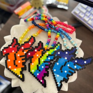 May include: A collection of colorful beaded necklaces with pixelated butterfly pendants. The pendants feature vibrant gradients of red, orange, yellow, green, blue, and purple. The necklaces are displayed on a white, flower-shaped dish.