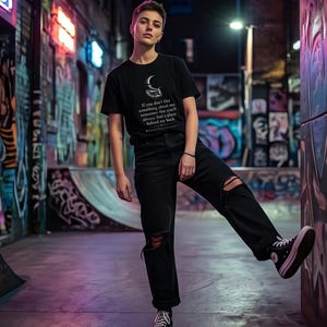 Può includere: Una t-shirt nera a maniche corte con la grafica di una luna crescente e il testo: If you don't like something about me, remember that you'll always find a place behind my back. Abbinata a jeans neri strappati e scarpe da ginnastica nere.