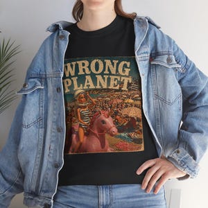 Puede incluir: Camiseta negra con un gráfico de estilo vintage que dice "WRONG PLANET". La imagen muestra a una mujer con un traje de baño a rayas y un casco montando un unicornio rosa en una playa. El gráfico tiene un estilo retro.