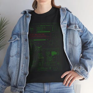Peut inclure: T-shirt noir avec un motif graphique vert et rouge rappelant une interface informatique. Le motif comprend du texte, une scène de plage et des lunettes de soleil. Le t-shirt est porté avec une veste en jean.