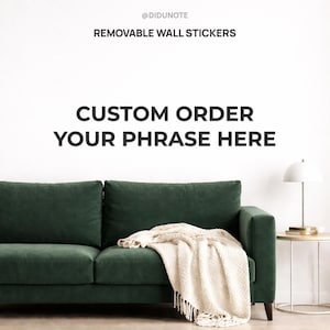 May include: A dark green velvet sofa with a cream throw. The wall features the text "CUSTOM ORDER YOUR PHRASE HERE" and "REMOVABLE WALL STICKERS". A white lamp is on a small side table.