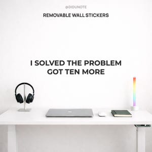 Op de afbeelding: Een witte bureau met een laptop, muis, notitieboek en koptelefoon. Rechts staat een kleurrijke LED-lamp. De tekst op de muur luidt "@DIDUNOTE REMOVABLE WALL STICKERS I SOLVED THE PROBLEM GOT TEN MORE."