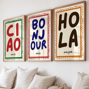 Op de afbeelding: Drie ingelijste kunstprints met de woorden "CIAO", "BONJOUR" en "HOLA" in vette letters. Elke print heeft een andere gekleurde rand en het woord "amici" of "amigos" onderaan. De prints hangen boven een houten bank met kussens.