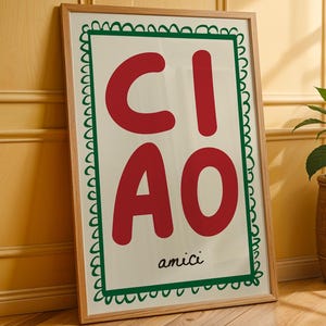 May include: A framed art print with the word "CIAO" in large red letters, and "amici" in small black letters. The artwork is framed in a light wood frame and has a green scalloped border. The print is leaning against a yellow wall.