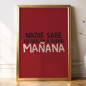 May include: A framed art print with a gold frame and a red background. The print features the Spanish text "NADIE SABE LO QUE VA A PASAR MAÑANA" in black and white. A decorative vase with dried plants is next to the frame.