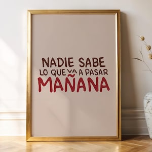 May include: A framed print with the Spanish text "NADIE SABE LO QUE VA A PASAR MAÑANA" in brown and red lettering. The frame is gold, and the background is a light beige color. A decorative vase with dried plants is to the right of the frame.