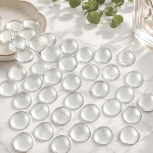 May include: A collection of numerous clear, circular glass cabochons with a smooth, domed surface. These transparent, pebble-like glass pieces are scattered across the surface, reflecting light and showcasing their uniform, rounded shape.