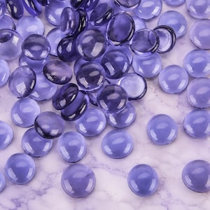 May include: A collection of numerous smooth, round glass gems in various shades of purple and violet. These translucent, circular decorative stones feature a polished finish and are scattered across the frame, displaying light reflections.