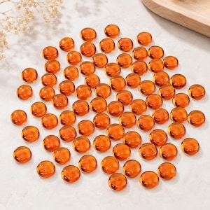 May include: A collection of numerous small, round, translucent amber-colored glass gems. Each piece features a smooth, domed top surface and a flat bottom, creating a uniform appearance across the entire set of decorative glass cabochons.
