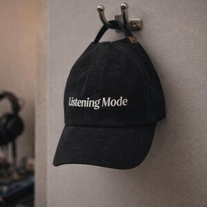 May include: Black baseball cap with the text "Listening Mode" in white. The cap is hanging on a silver hook on a grey wall. The background shows a recording studio with audio equipment.