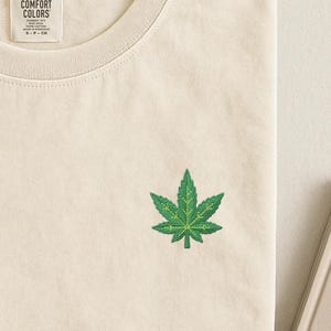 Puede incluir: Una camiseta de color crema con un pequeño diseño de hoja de cannabis bordado en verde en el pecho. La camiseta tiene un cuello redondo acanalado y una etiqueta interior blanca que dice COMFORT COLORS, GARMENT DYE, RING SPUN, 100% COTTON, MADE IN HONDURAS, S-P-CH.