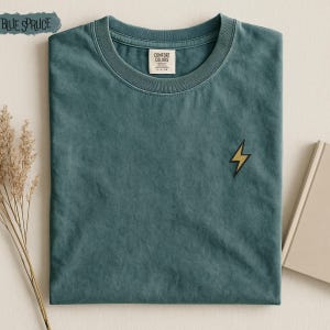 May include: A blue spruce colored short-sleeve t-shirt featuring a small embroidered yellow lightning bolt on the chest. The shirt has a crew neckline and a Comfort Colors brand label on the inside collar. The fabric has a washed, matte finish.