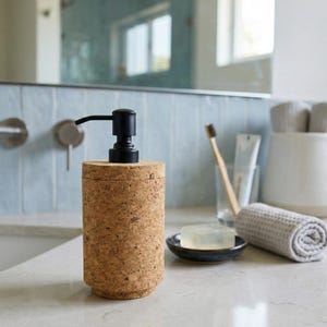 Puede incluir: Dispensador de jabón cilíndrico hecho de corcho natural con una bomba negra. El dispensador está sobre una encimera blanca, con una toalla gris enrollada, jabón y un cepillo de dientes en el fondo. El baño tiene una pared de azulejos azul claro y un espejo.
