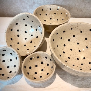 Può includere: Un set di cinque ciotole in ceramica di varie dimensioni. Le ciotole sono bianco sporco con finitura maculata e pois neri distanziati. Le ciotole sono disposte su una superficie bianca.