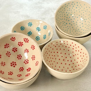 Könnte beinhalten: Eine Sammlung kleiner, cremefarbener Keramikschalen. Einige Schalen weisen rote Blumenmuster auf, andere blaue Blumendesigns und einige blaue Punkte. Die Schalen sind gestapelt und zeigen ihre dekorativen Innenräume.