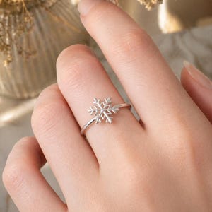 Puede incluir: Un anillo de copo de nieve plateado. El anillo presenta un delicado diseño de copo de nieve como pieza central, con detalles intrincados. El anillo se muestra en un dedo, destacando su tamaño y diseño. Es una joya.