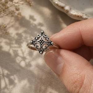 Puede incluir: Un anillo de plata con un diseño ornamentado y simétrico. El centro del anillo presenta un patrón en forma de diamante con detalles en espiral y pequeños acentos circulares. El anillo es sostenido por una mano, sobre un fondo suave y neutro.