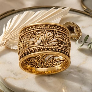 Può includere: Un bracciale a polsino color oro con intricati disegni floreali e a volute. L'ampia fascia presenta motivi dettagliati e una superficie strutturata. Il bracciale è esposto su una superficie di marmo con una foglia di palma secca e una geode.