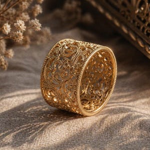 May include: A gold ring with an elaborate filigree design. The ring features a wide band adorned with detailed floral and scroll patterns. It is displayed on a neutral fabric, with soft lighting enhancing the intricate details of the ring.