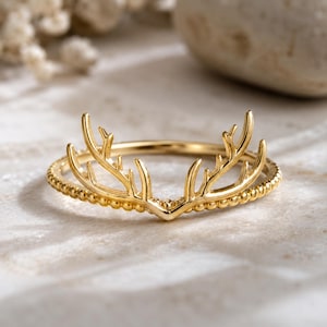 May include: A gold-colored ring featuring a detailed antler design. The ring has a beaded band and the antlers are the focal point, with intricate detailing. The ring is set against a neutral background, highlighting the jewelry's design.