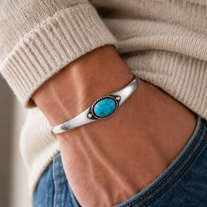 Pode incluir: Uma pulseira de prata tipo bracelete com uma pedra turquesa oval central com veios escuros. A pedra está engastada numa borda de corda torcida e ladeada por duas pequenas esferas de prata numa banda de prata lisa e polida que se estreita.