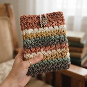 May include: A rectangular crochet pouch featuring horizontal stripes in shades of terracotta, cream, mustard yellow, sage green, and dark gray. The pouch is secured with a light blue cord and a wooden charm shaped like a leafy branch.
