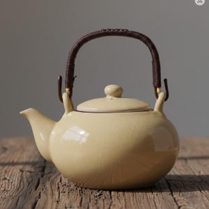 May include: A cream-colored ceramic teapot with a brown woven handle and lid. The teapot has a rounded body, a curved spout, and a small knob on the lid. The handle is attached with metal hardware.