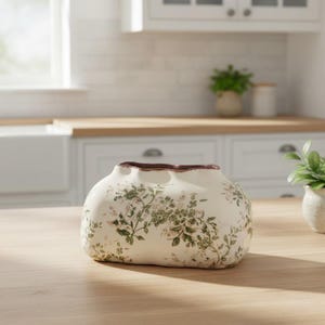 Pode incluir: Vaso de cerâmica creme com um padrão floral em verde e rosa. O vaso tem uma borda superior ondulada distinta com uma borda marrom. Ele está em uma superfície de madeira clara, adequado para decoração.