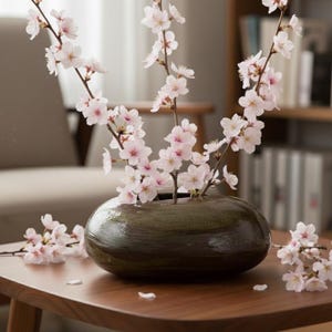 Può includere: Un vaso di ceramica verde scuro, di forma ovale, contiene rami di fiori di ciliegio rosa. Il vaso è su un tavolo di legno, con altri fiori sparsi. La scena evoca la primavera e la bellezza naturale.